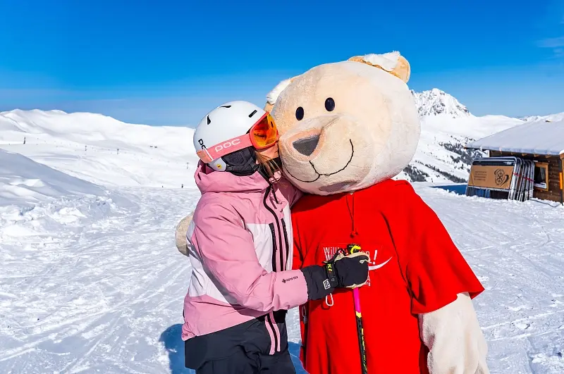 A skier in a helmet hugging the Wildkogel-Arena bear mascot on the piste