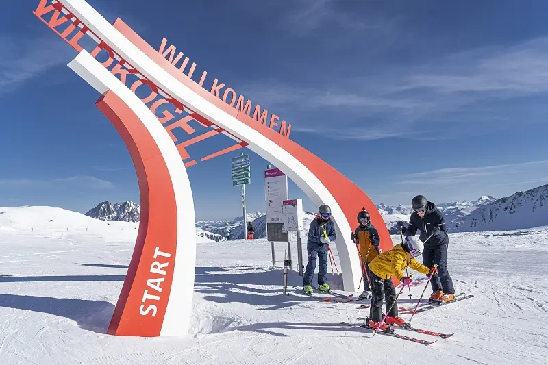 A family on skis under the red “Willkommen Wildkogel” start arch on the plateau, with the snow-covered peaks of the Hohe Tauern behind